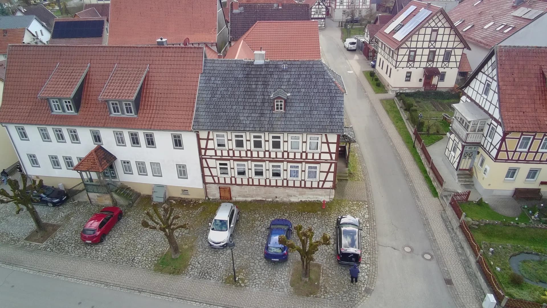 Vogelperspektive auf das historische Gebäudeensemble im Ortskern mit Fachwerkhäusern, gepflastertem Vorplatz/Parkbereich und altem Baumbestand.