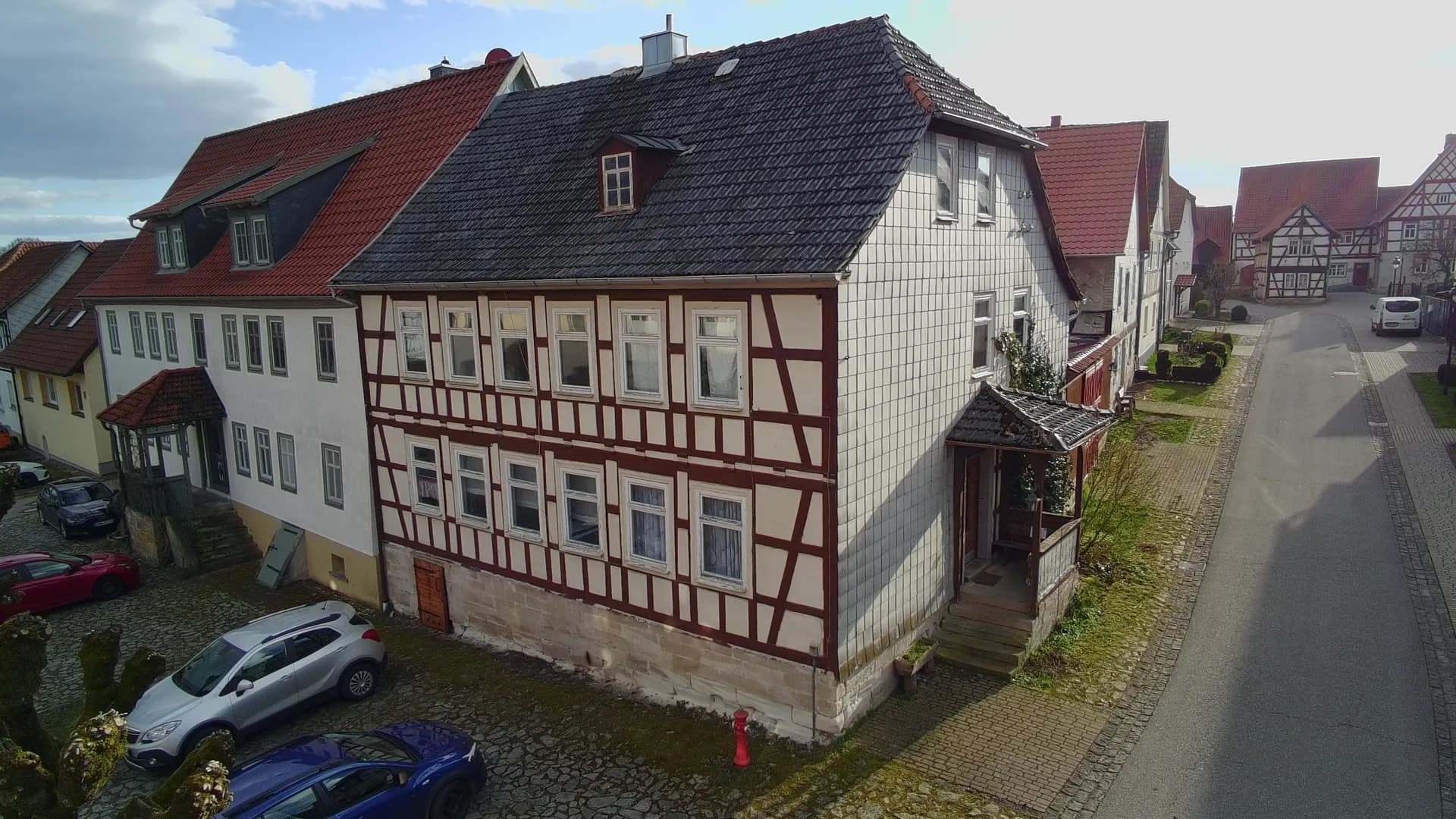 Perspektivische Eckansicht des Fachwerkhauses mit schieferverkleideter Giebelseite, überdachtem Treppeneingang und Blick in die historische Dorfstraße.