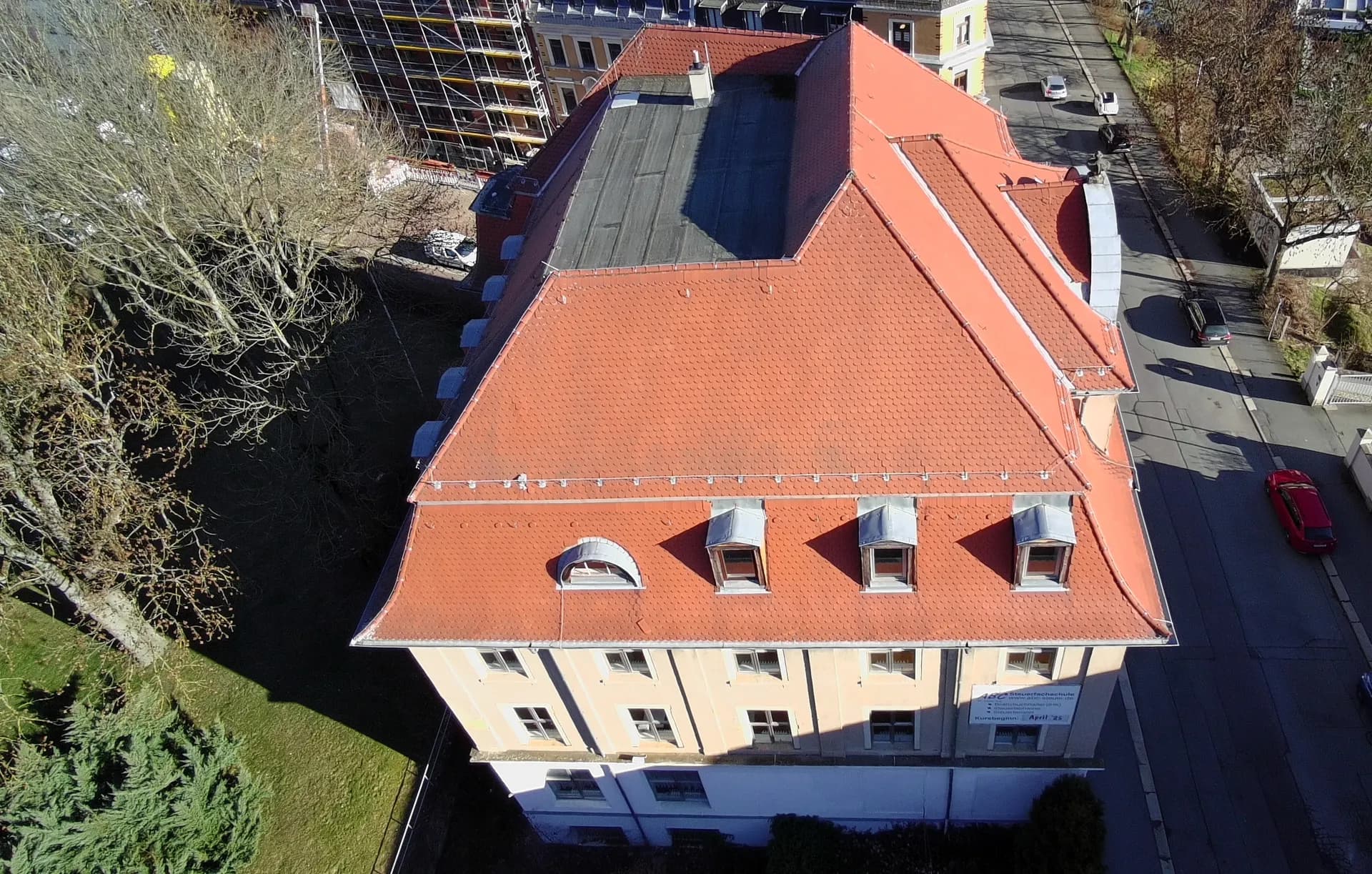 Drohnenaufnahme der markanten Mansarddachkonstruktion mit roten Tonziegeln, stehenden Zinkblechgauben und Blick auf den Straßenraum.