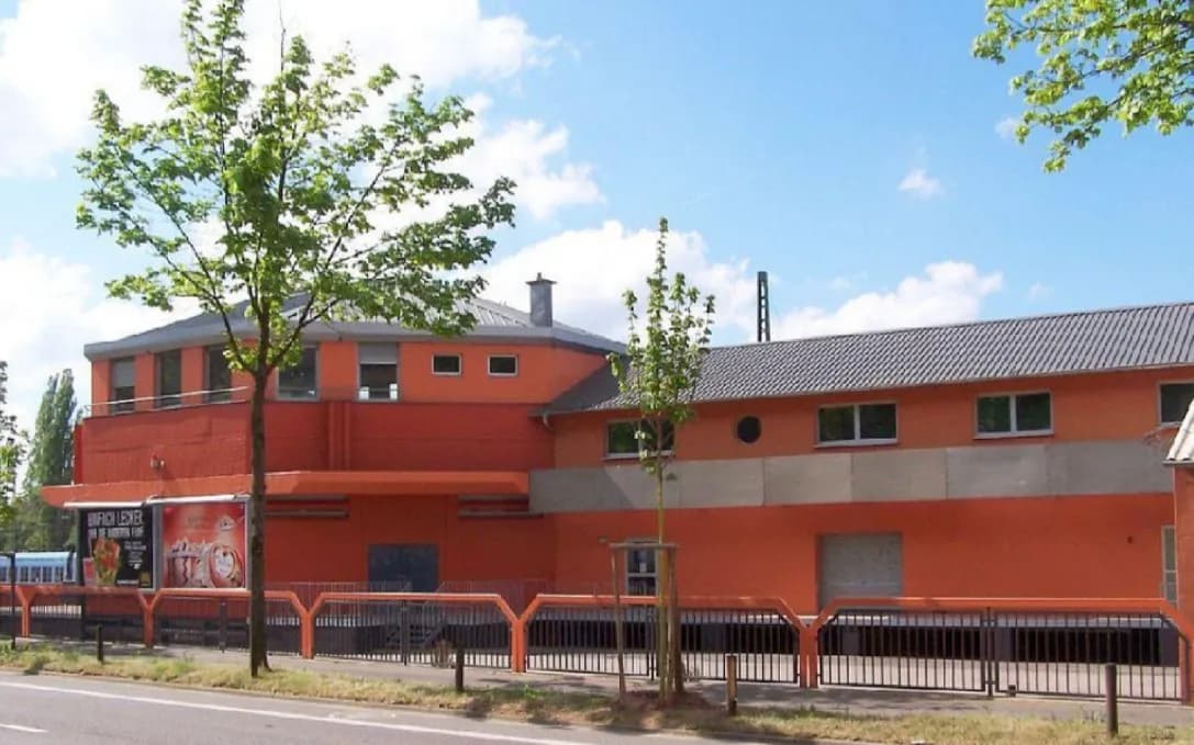Straßenansicht der Gewerbe- und Gastronomieimmobilie in markantem Terrakotta-Orange mit polygonalem Kopfbau, Blechdach, Hallentrakt und eingefriedetem Vorplatz.  Screenshot 2026-03-14 065023.jpg