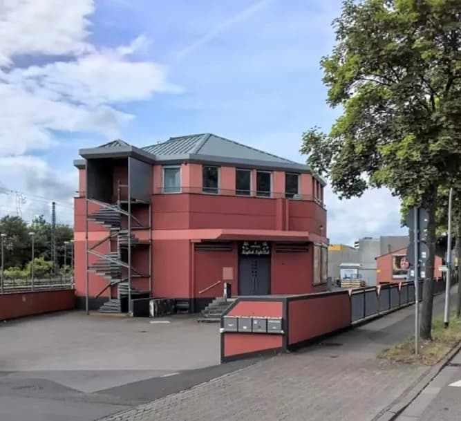 Außenansicht des Event-/Clubgebäudes mit Haupteingang, stählerner Wendeltreppe als Fluchtweg, großzügigem asphaltiertem Vorplatz und integrierter Briefkastenanlage.