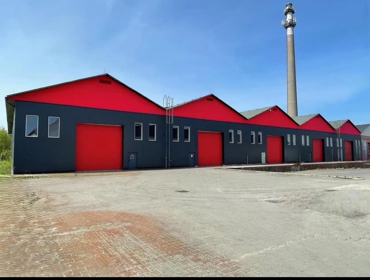 Moderne Gewerbe- und Lagerhalle mit markanter rot-schwarzer Fassade, mehreren Sektionaltoren, Antennenmast und großzügiger gepflasterter Hoffläche zur Anfahrt.