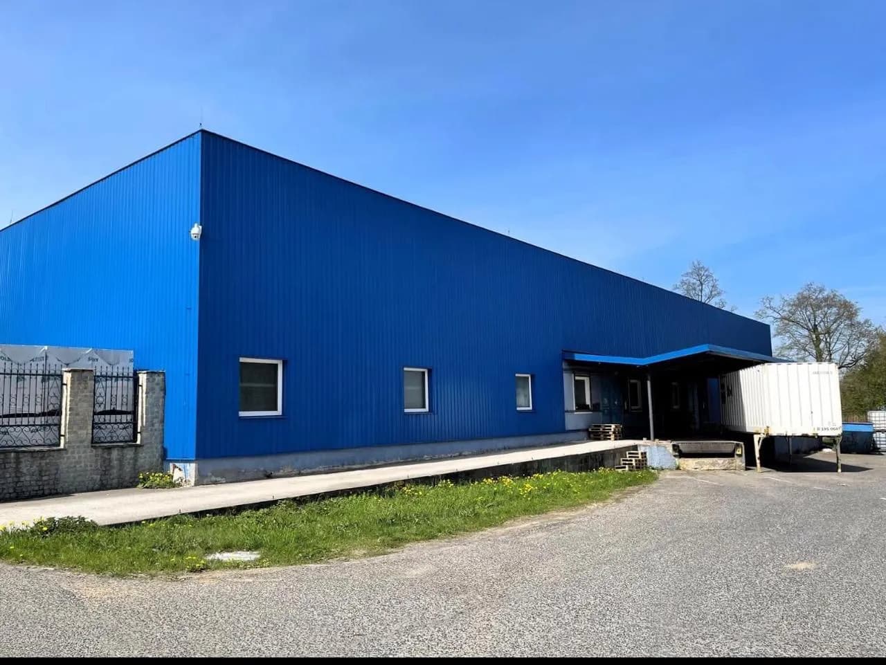 Außenansicht einer blauen Lager- oder Logistikhalle mit Paneelverkleidung, Laderampe samt Überdachung und befahrbarem Außenbereich.
