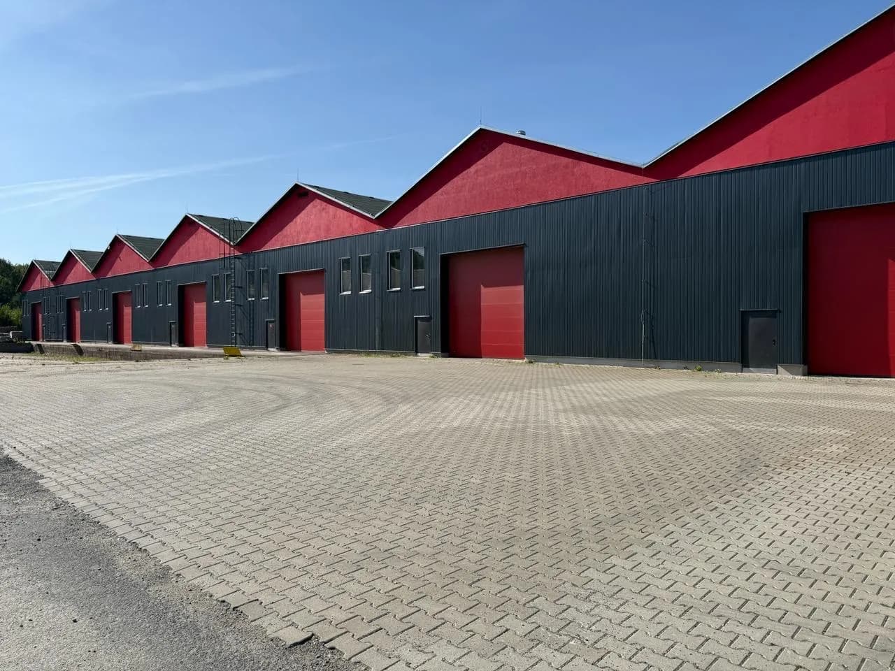 Fassade der Gewerbehalle mit markantem Sheddach, rot-schwarzer Verkleidung, mehreren ebenerdigen Toren und gepflasterter Rangierfläche.