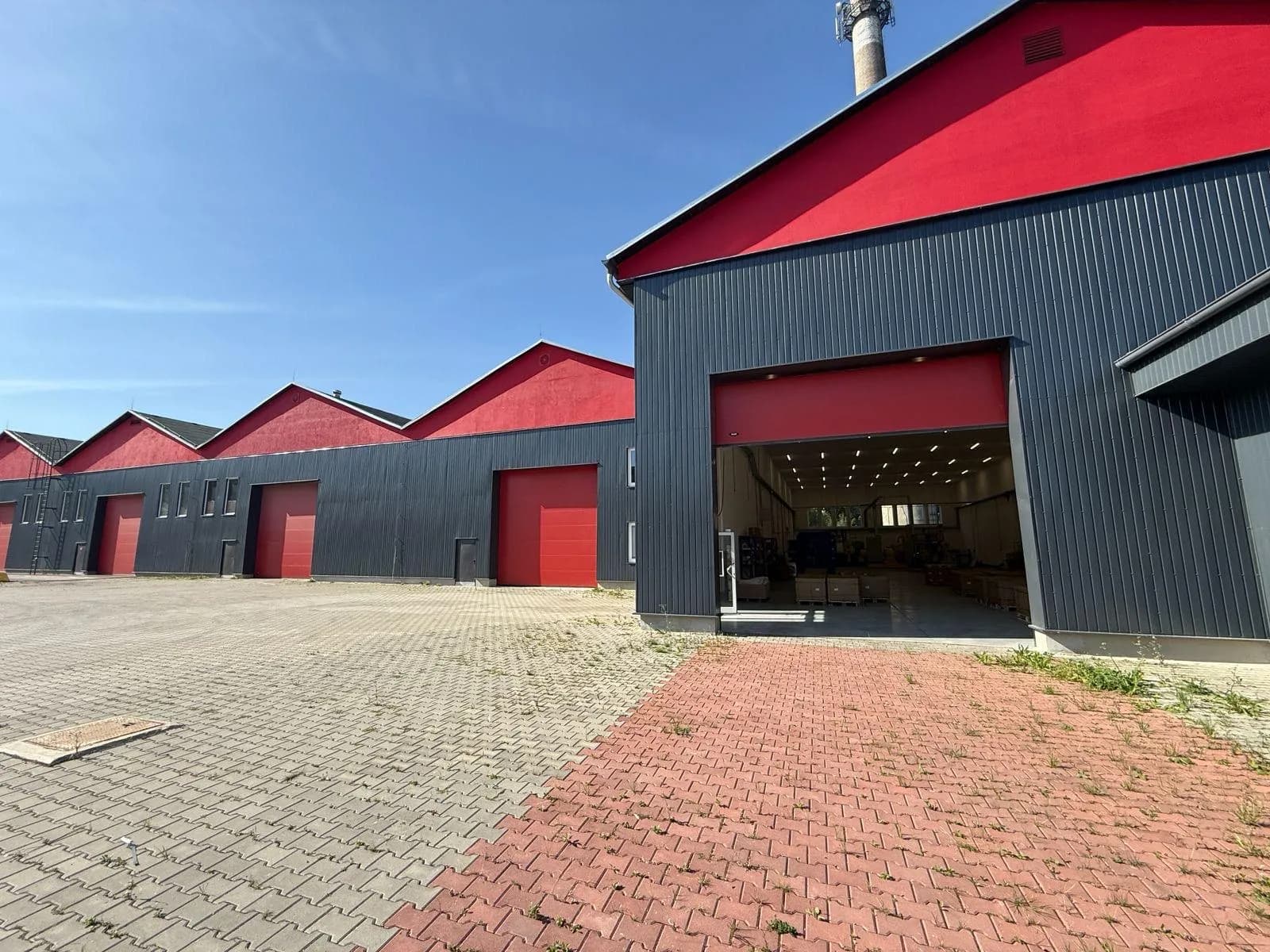 Außenansicht des Gewerbekomplexes mit Sheddach, geöffnetem Sektionaltor zur beleuchteten Halle und großzügigem gepflastertem Rangier- und Vorplatz.