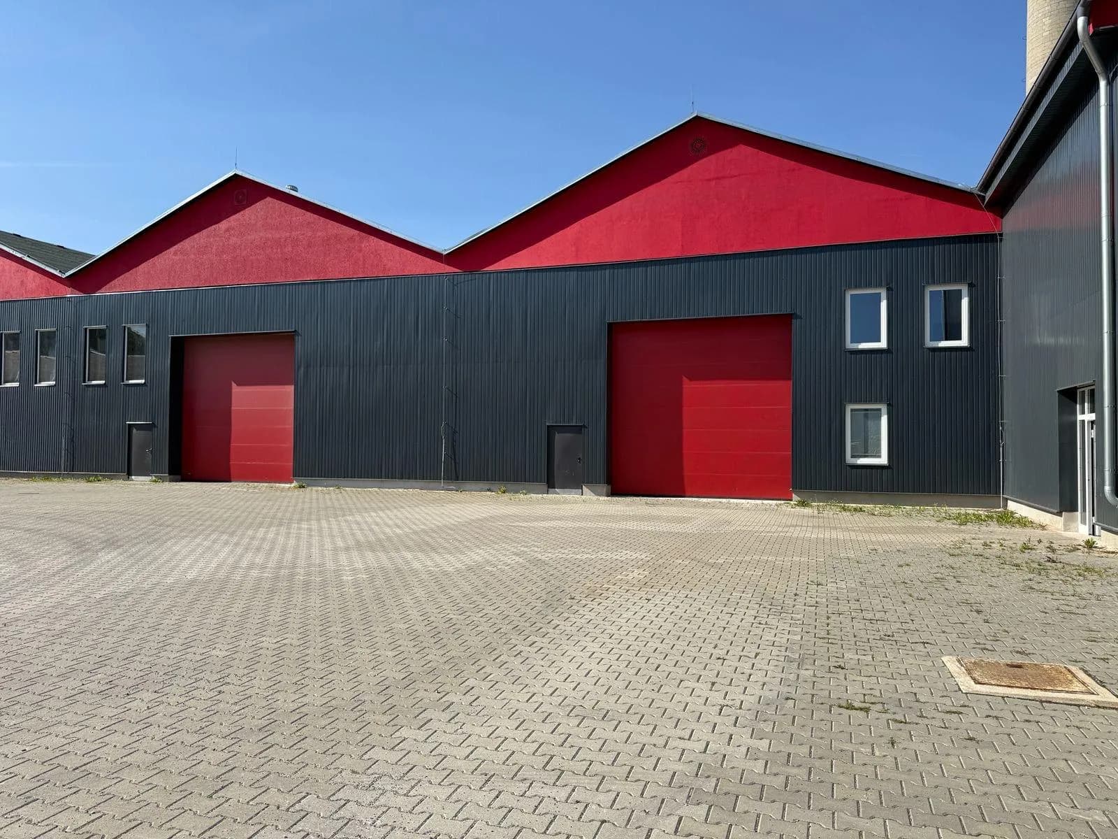 Frontalansicht der Gewerbehalle mit roten Sektionaltoren, vertikaler Fassadenverkleidung und weitläufig gepflasterter Vor- und Rangierfläche für Nutzfahrzeuge.