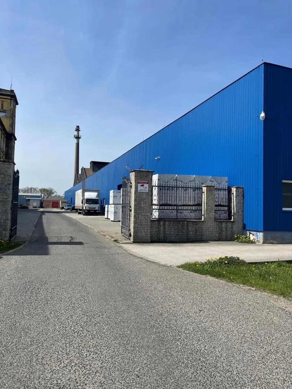 Zufahrt zum umzäunten Gewerbegelände mit blauer Lagerhalle, Toranlage und befestigter Anfahrtsstraße für Lkw und Lieferverkehr.