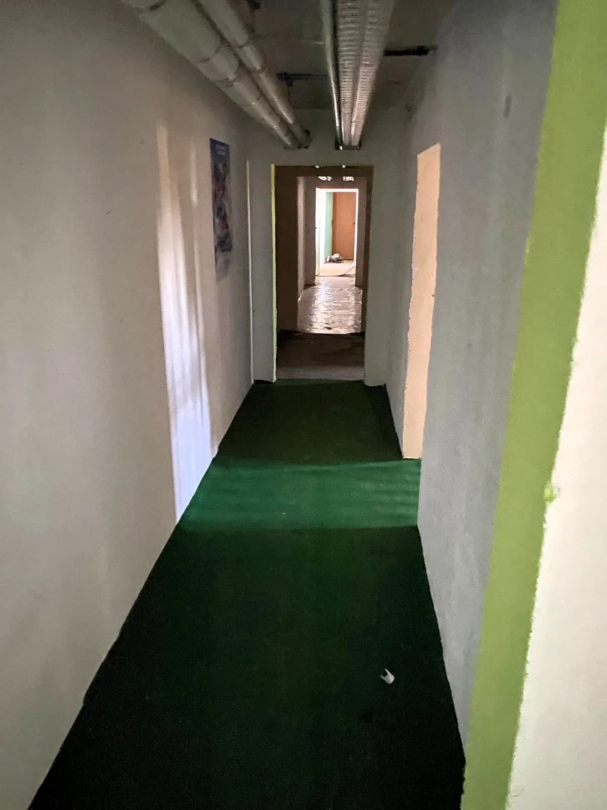 Langer Wohngebäudeflur mit grünem Bodenbelag, hellen Wänden, freiliegenden Deckenrohren und Durchgängen zu den einzelnen Wohneinheiten.