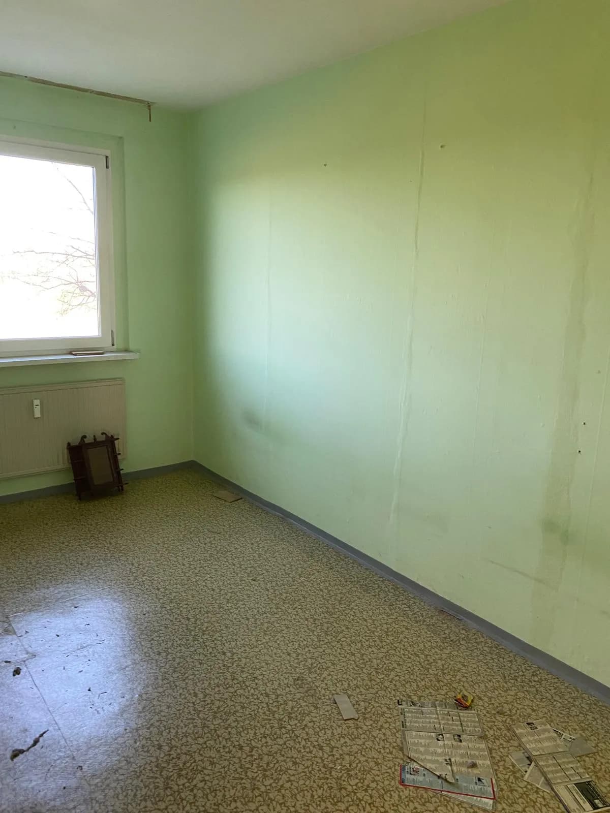 Renovierungsbedürftiges, helles Zimmer im Plattenbau mit blassgrünen Wänden, Isolierglasfenster, Heizkörper und gemustertem Linoleumboden