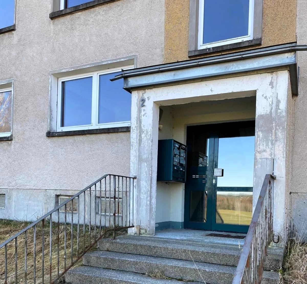 Eingangsbereich eines mehrstöckigen Wohngebäudes vor der Sanierung: Betontreppe mit Metallgeländer, verglaste Hauseingangstür, Vordach und Briefkastenanlage an der Fassade.