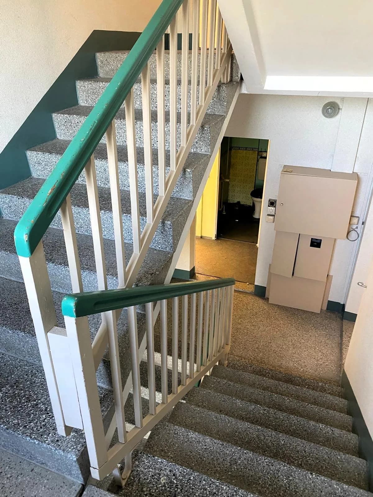 Helles Treppenhaus im Plattenbau: Terrazzostufen, klassisches Geländer mit türkisfarbenem Handlauf und Stromverteilerkästen auf dem Zwischenpodest
