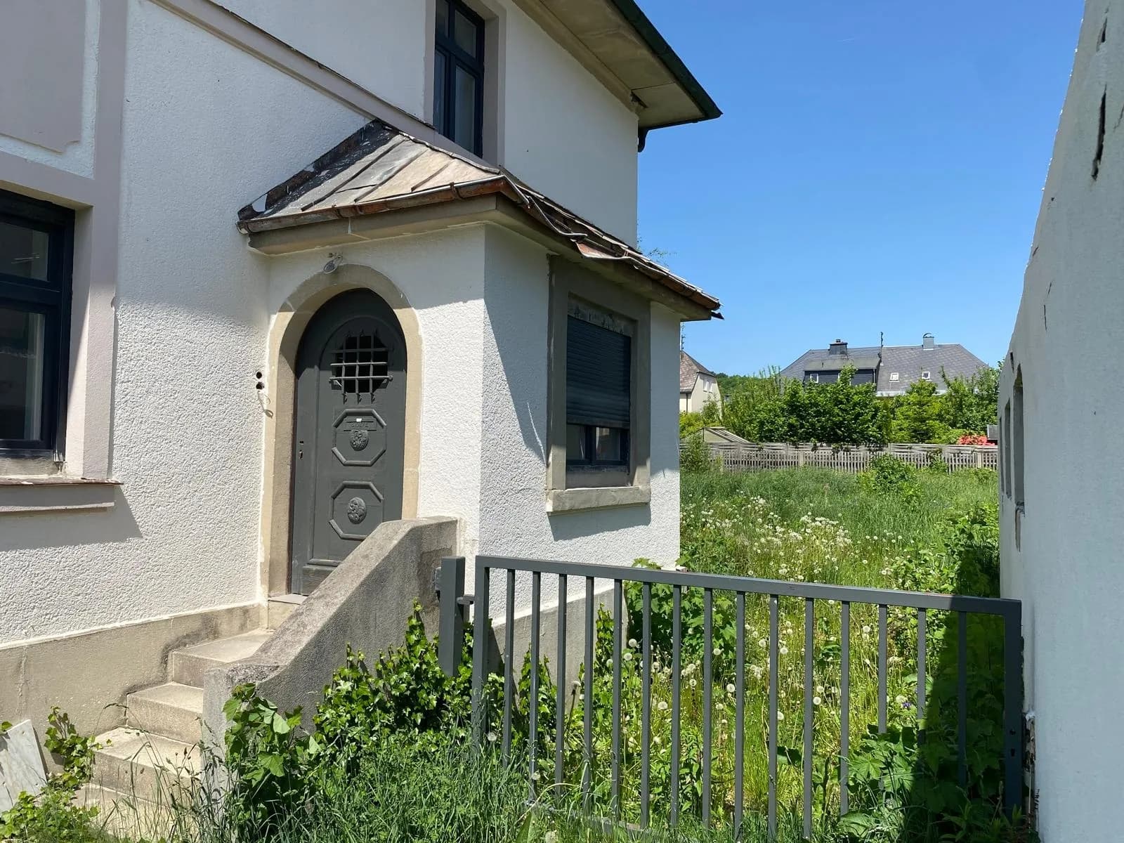 Seitlicher Hauseingang mit Betontreppe, gewölbter grauer Haustür, Metallzaun und Blick in den begrünten Gartenbereich.