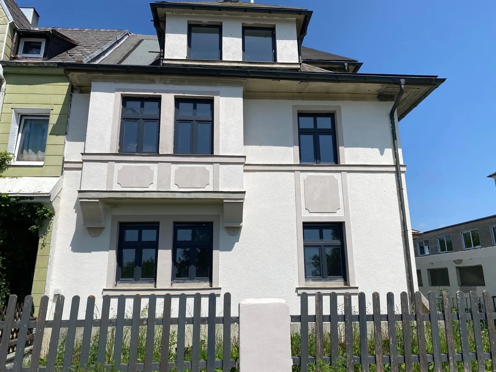 Helle Hausfassade des Mehrfamilienhauses mit Erker, dunklen Fensterrahmen, Dachgaube und Holzzaun im Vordergrund.
