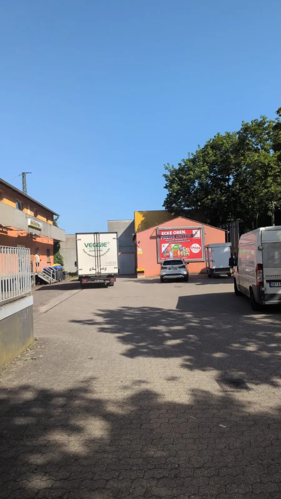 Asphaltierter Gewerbe- und Wirtschaftshof mit Pkw-Stellplätzen, Anlieferungszone für Lieferwagen und Lkw, Nebengebäuden und Werbetafel an der Fassade.