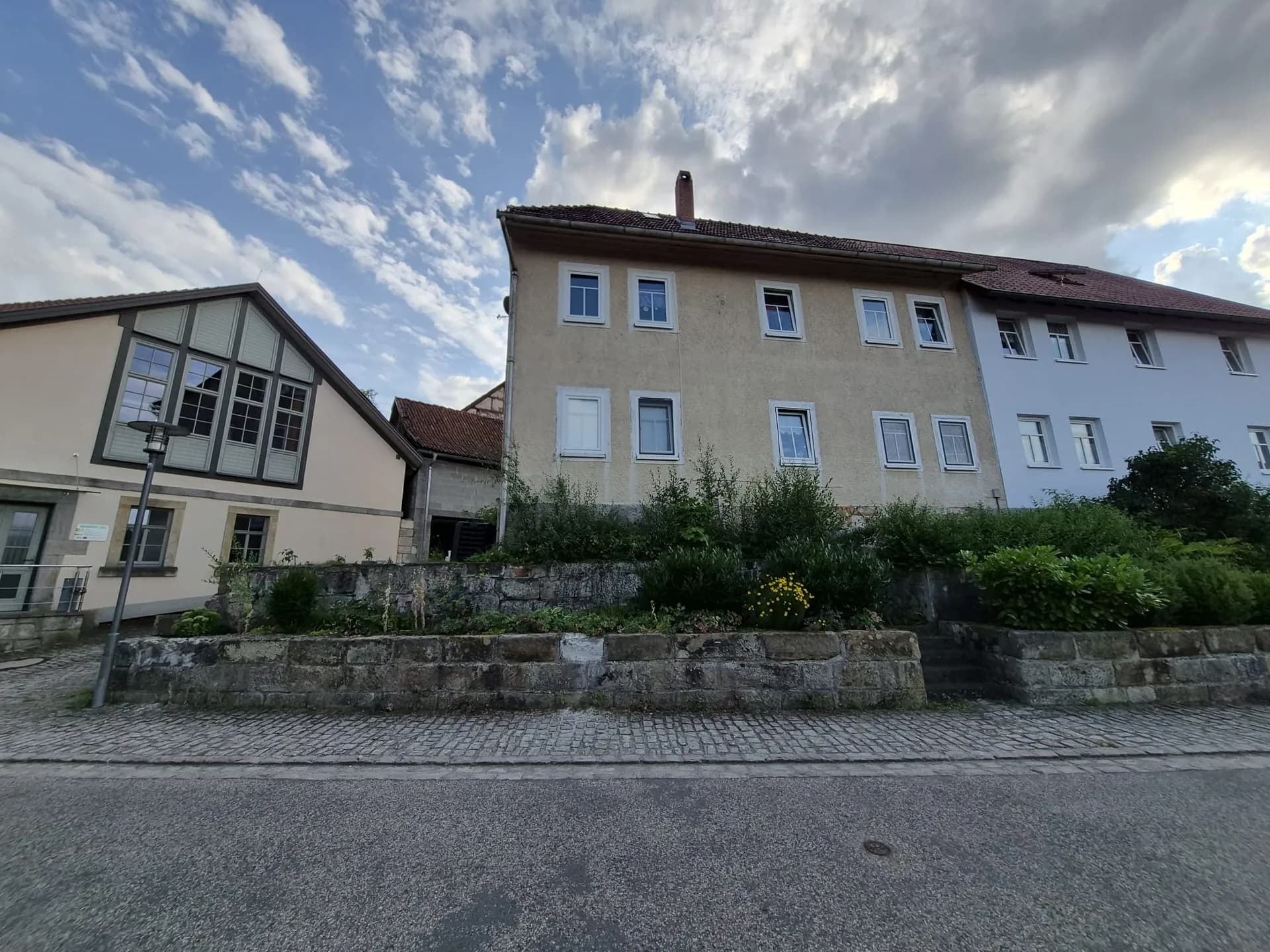 Straßenansicht des Mehrfamilien-/Wohnhauses mit erhöhtem Vorgartenbereich, Naturstein-Stützmauer und angrenzenden Gebäudeteilen.