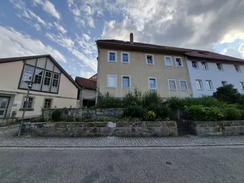 Straßenansicht des Mehrfamilien-/Wohnhauses mit erhöhtem Vorgartenbereich, Naturstein-Stützmauer und angrenzenden Gebäudeteilen.
