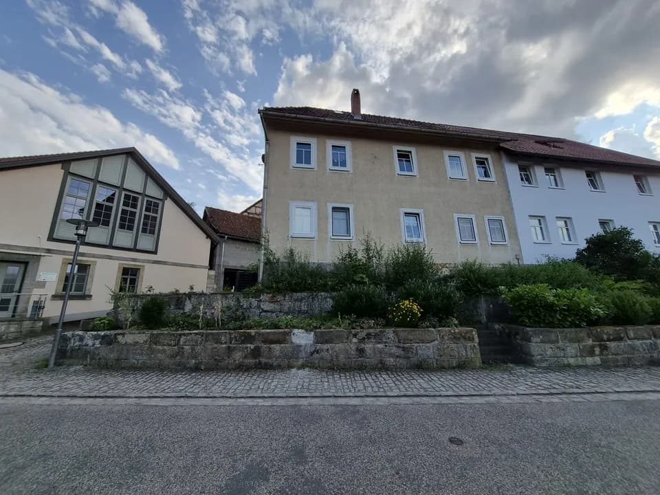 Straßenansicht des Mehrfamilien-/Wohnhauses mit erhöhtem Vorgartenbereich, Naturstein-Stützmauer und angrenzenden Gebäudeteilen.