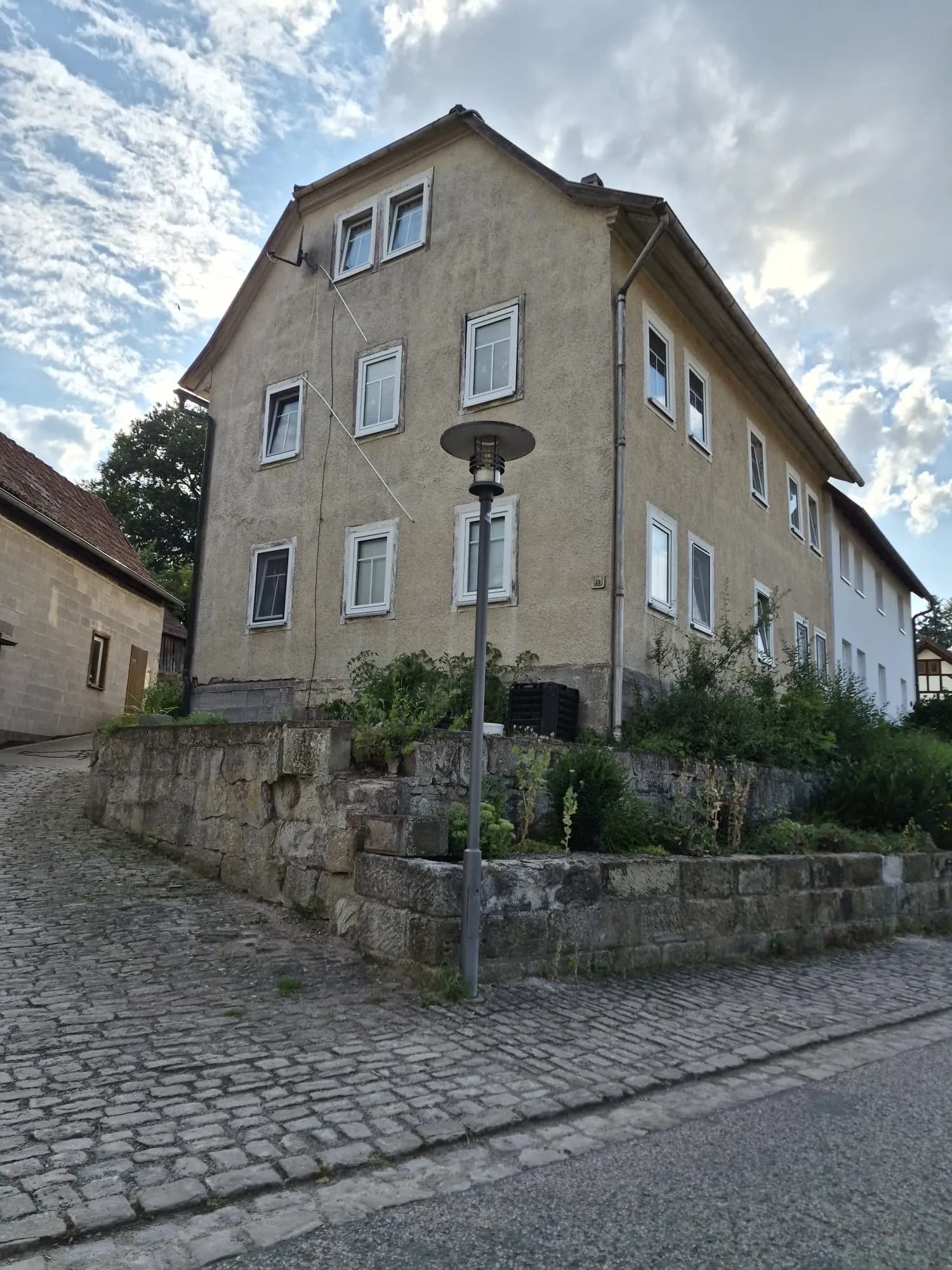 Eckansicht des massiven Wohnhauses auf erhöhtem Natursteinsockel an einer verkehrsberuhigten, gepflasterten Straße.