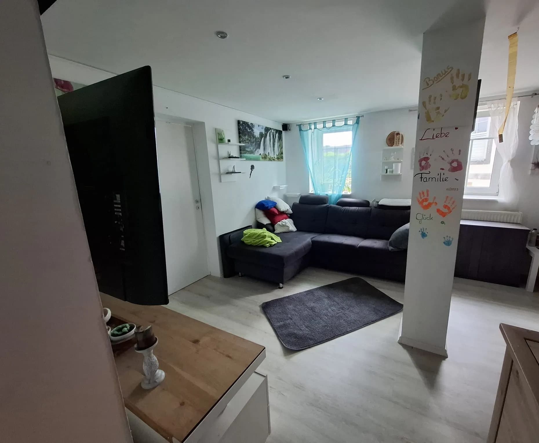 Helles Wohnzimmer mit dunkler Eckcouch, Teppich, Flachbild-TV, Tageslichtfenster und modernem hellen Laminatboden.