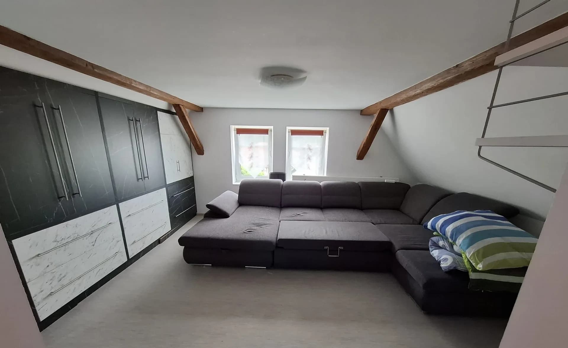Gemütlicher Wohnbereich im Dachgeschoss mit sichtbaren Holzbalken, großer Wohnlandschaft, Einbauschränken mit modernen Fronten und Fenstern.