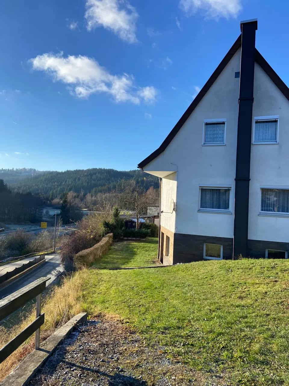 Seitenansicht des Wohnhauses mit ansteigendem Gartengrundstück, Außenkamin und idyllischem Fernblick auf Wald und Berge.
