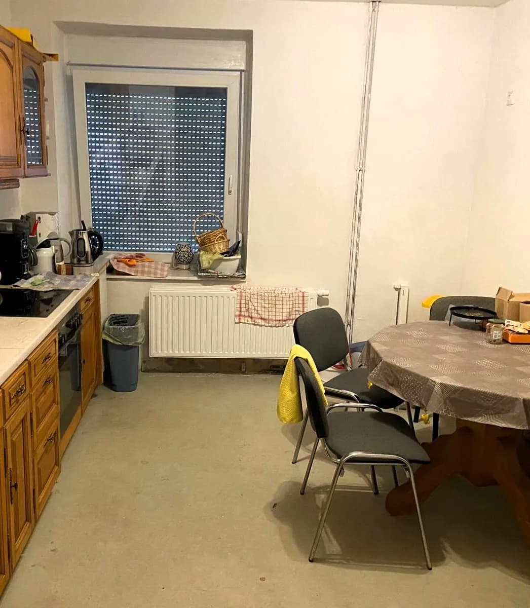 Geräumige Wohnküche mit Holz-Einbauzeile, Kochfeld, Backofen, rundem Esstisch und Kunststofffenster samt Außenrollladen.