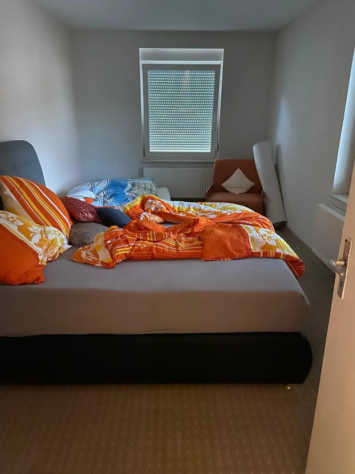 Schlafzimmer mit großem Doppelbett, Polstersessel, Teppichboden und Tageslichtfenster inklusive Außenrollladen.