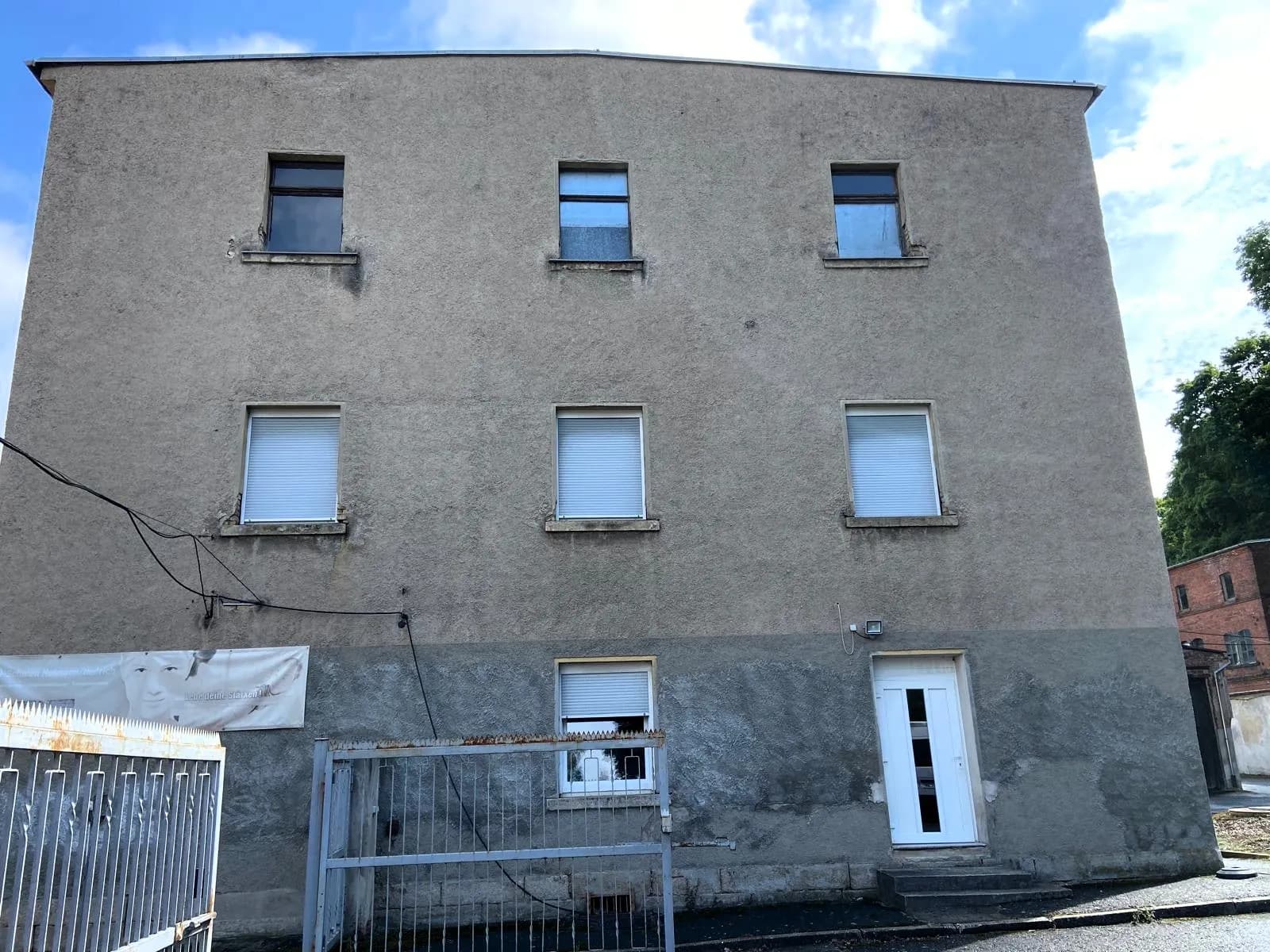Straßenseitige Giebelfassade des dreigeschossigen Gebäudes mit Putzoberfläche, weißer Hauseingangstür samt Stufen, Rollladenfenstern und metallenem Hoftor.