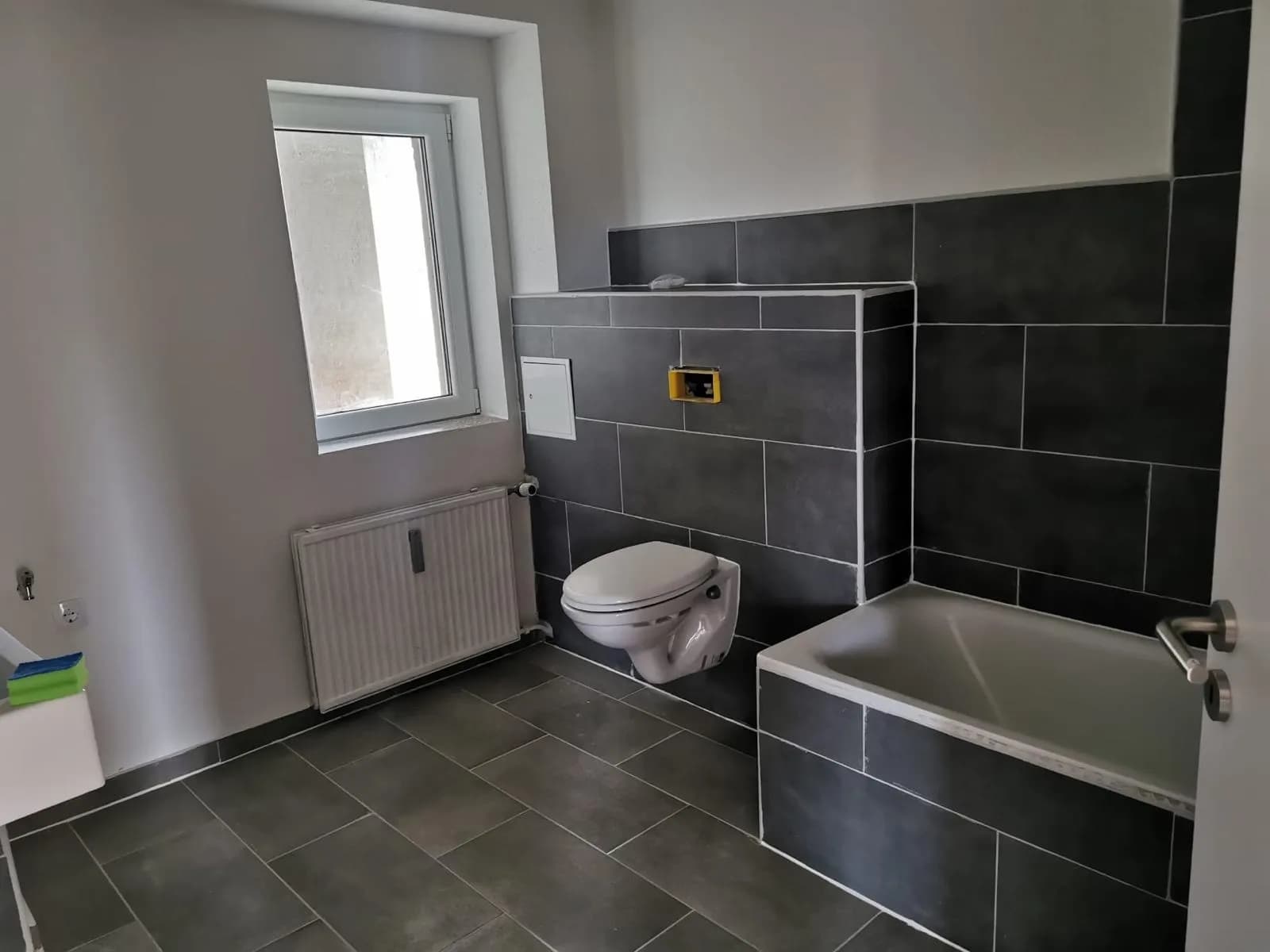Modern saniertes Badezimmer mit dunkelgrauen Großformatfliesen, Einbaubadewanne, wandhängendem WC mit Vorwandinstallation, Heizkörper und Tageslichtfenster.