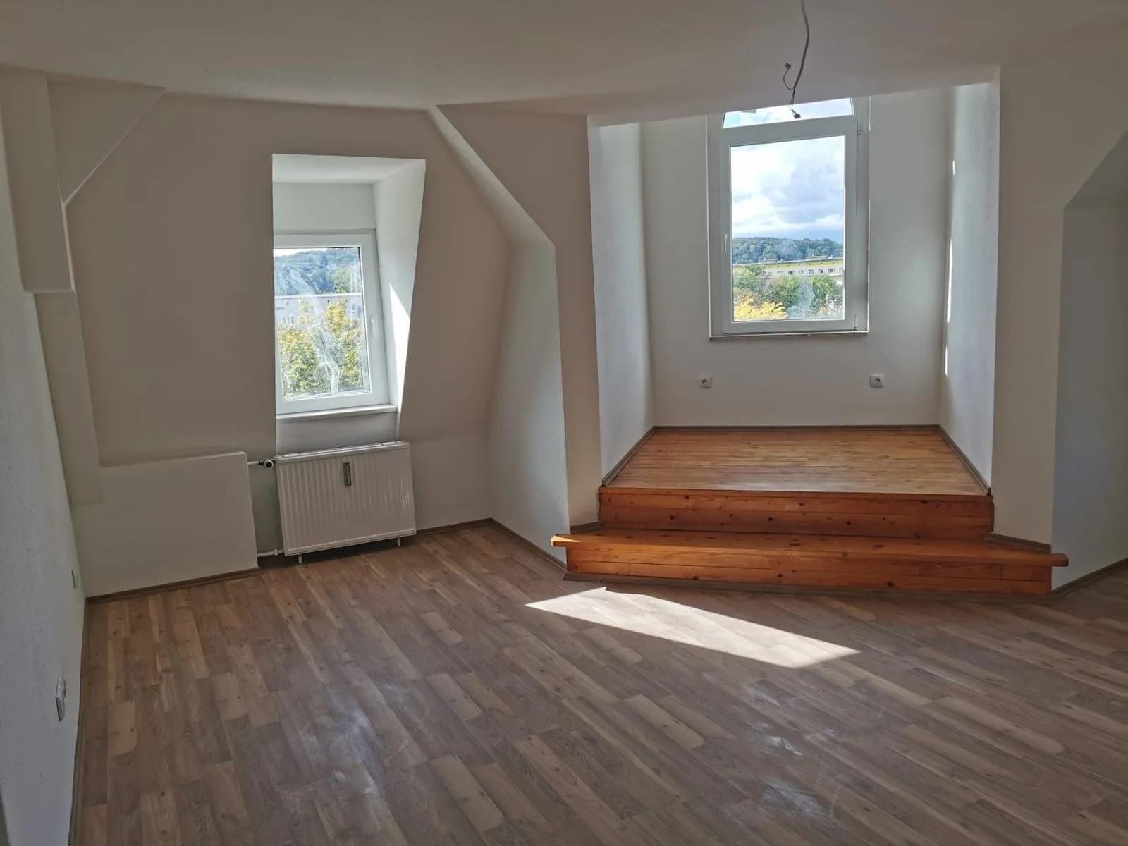 Großzügiges Dachgeschosszimmer mit Laminatboden, Heizkörper, zwei Fenstern und einer charmant erhöhten Holzpodest-Ebene im Gaubenbereich.