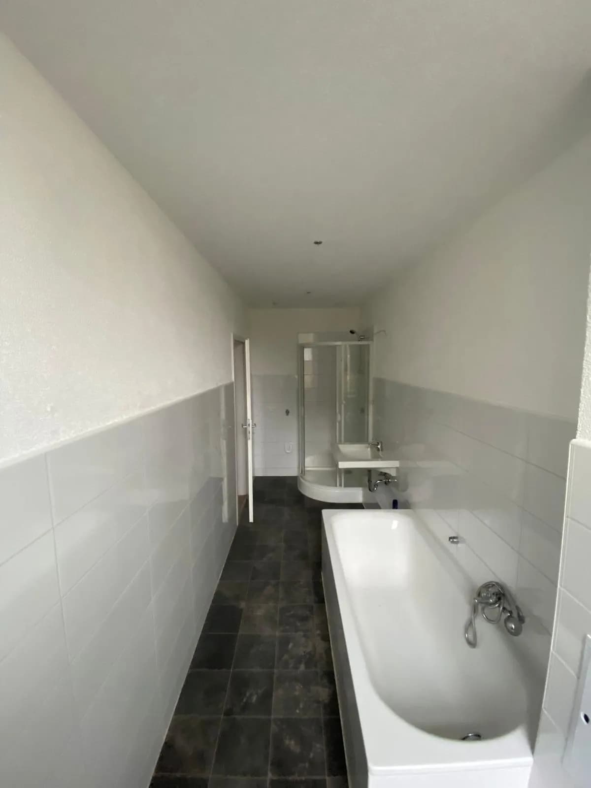 Längliches, helles Badezimmer mit Badewanne, separater Runddusche, Waschtisch, weißen Wandfliesen und dunklem Fliesenboden.