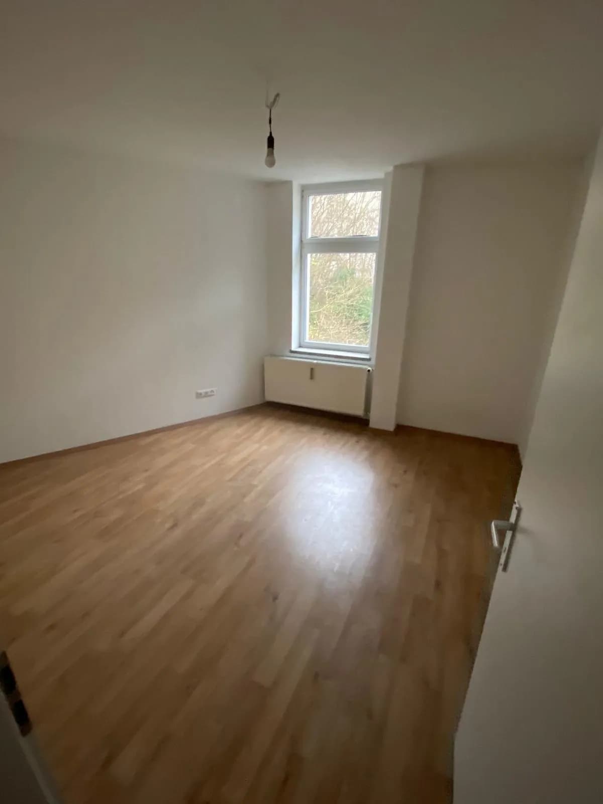 Helles Schlafzimmer mit vorteilhaftem Schnitt, gepflegtem Laminatboden, Heizkörper und hohem Fenster mit Ausblick ins Grüne.