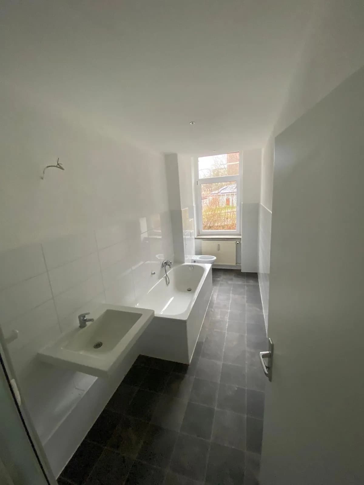 Tageslichtbad mit dunklem Fliesenboden, weißem Waschtisch, Einbaubadewanne und WC-Bereich am Fenster.