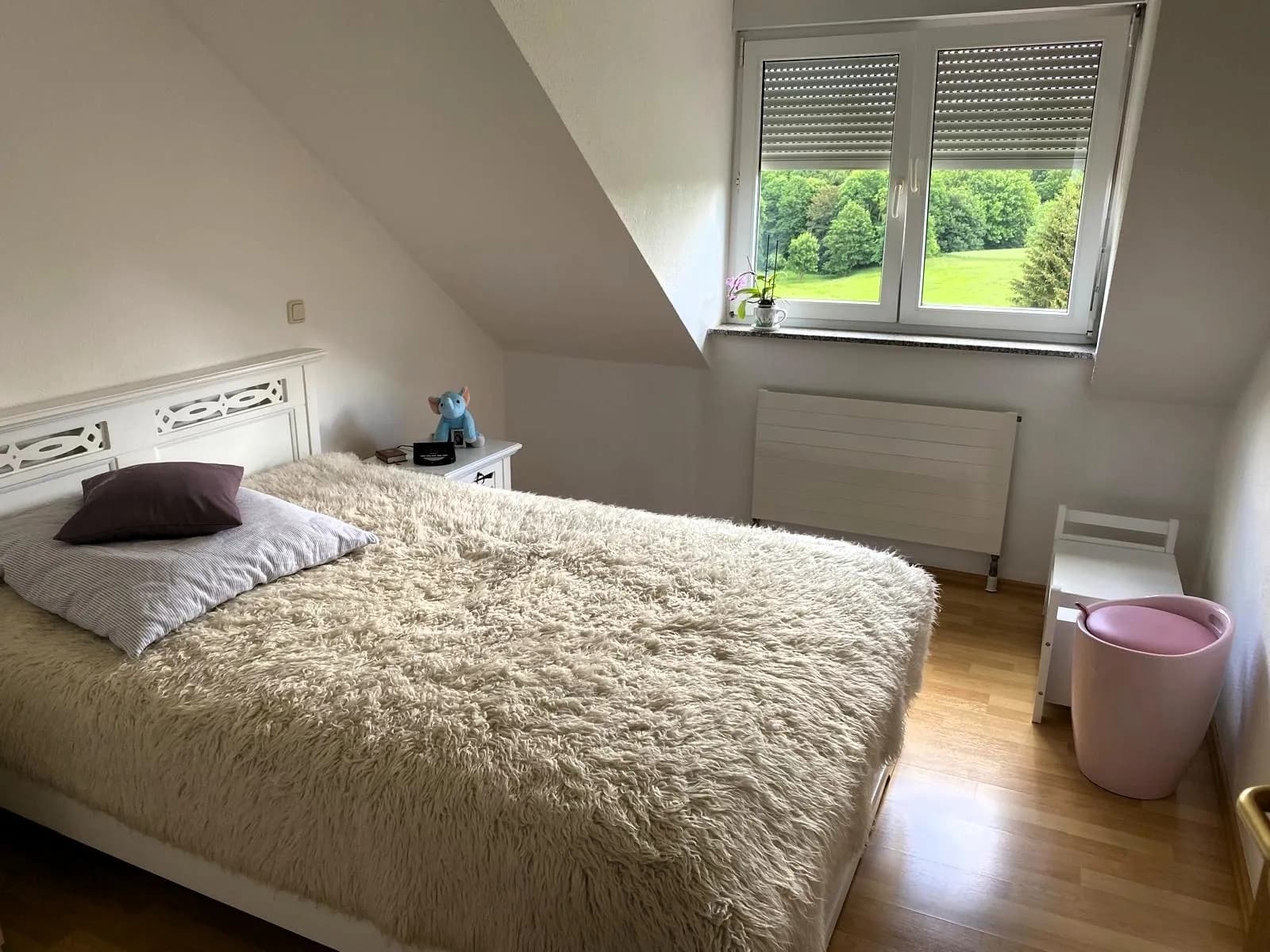 Gemütliches Schlafzimmer im Dachgeschoss mit Wandschräge, weißem Doppelbett, Heizkörper und Rollladenfenster mit idyllischem Blick auf Wiesen und Wald.