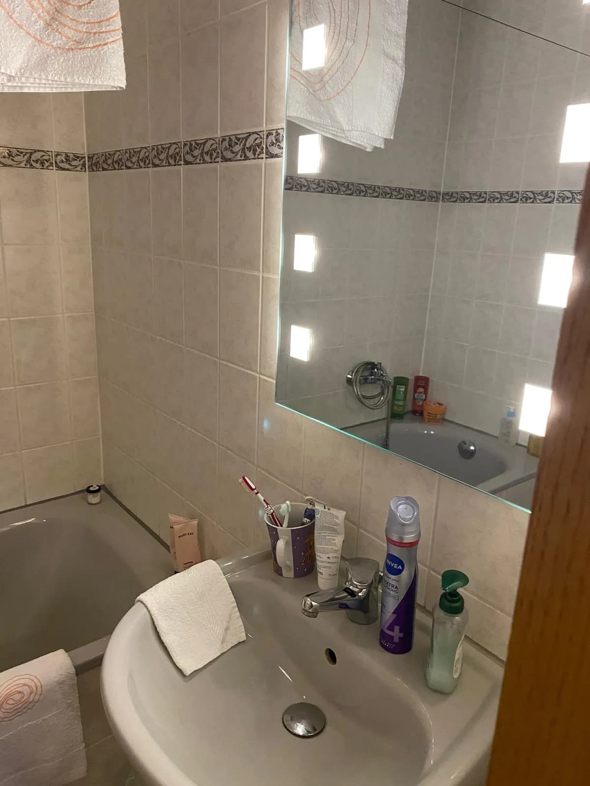 Badezimmer mit beigefarbenen Fliesen und Zierbordüre, Einbaubadewanne, Waschtischarmatur und großem Wandspiegel mit integrierter Beleuchtung.