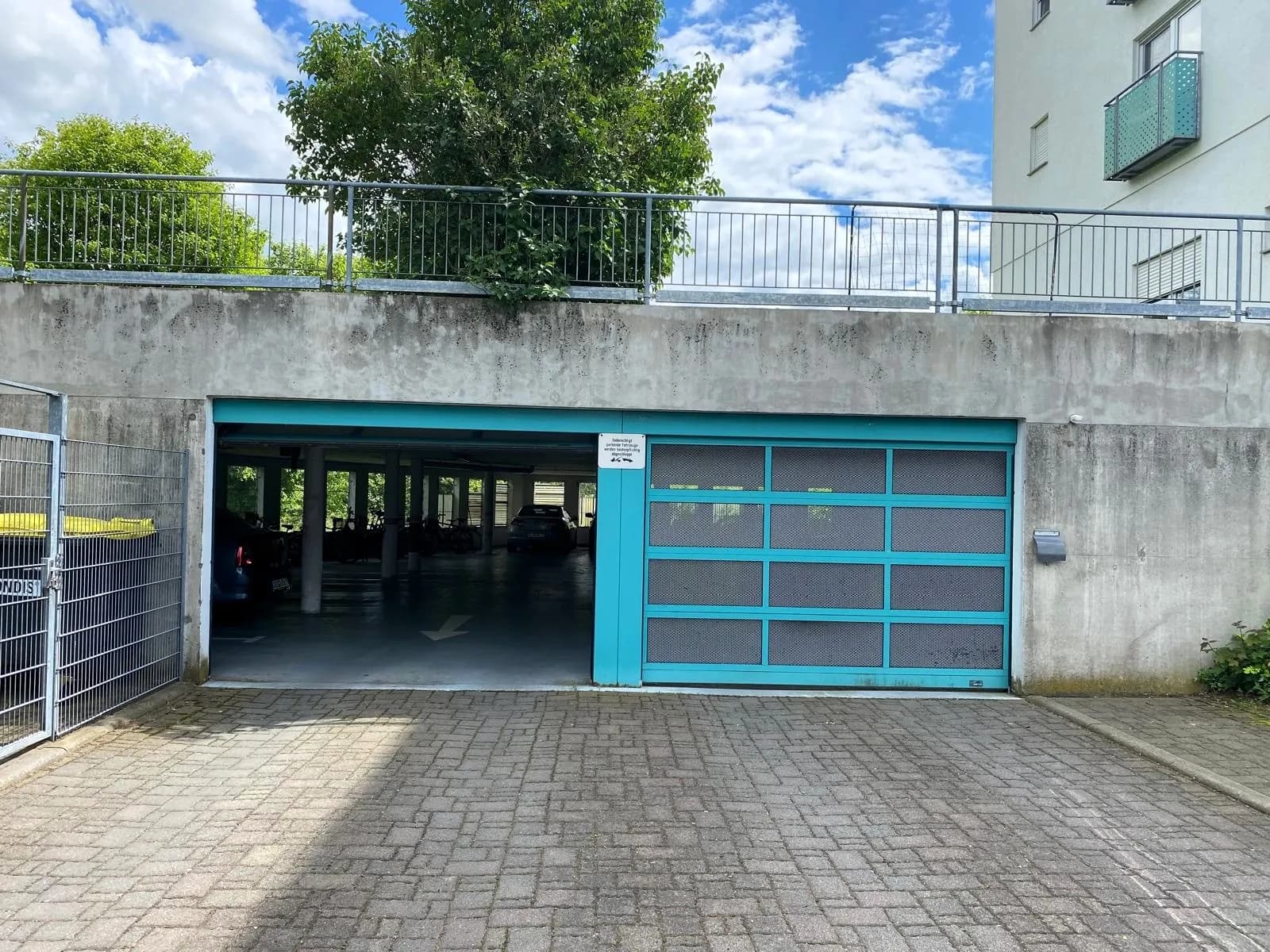 Einfahrt zur Tiefgarage mit türkisfarbenem Gitter-Rolltor, gepflastertem Vorplatz und oberhalb liegender Terrassenbrüstung.