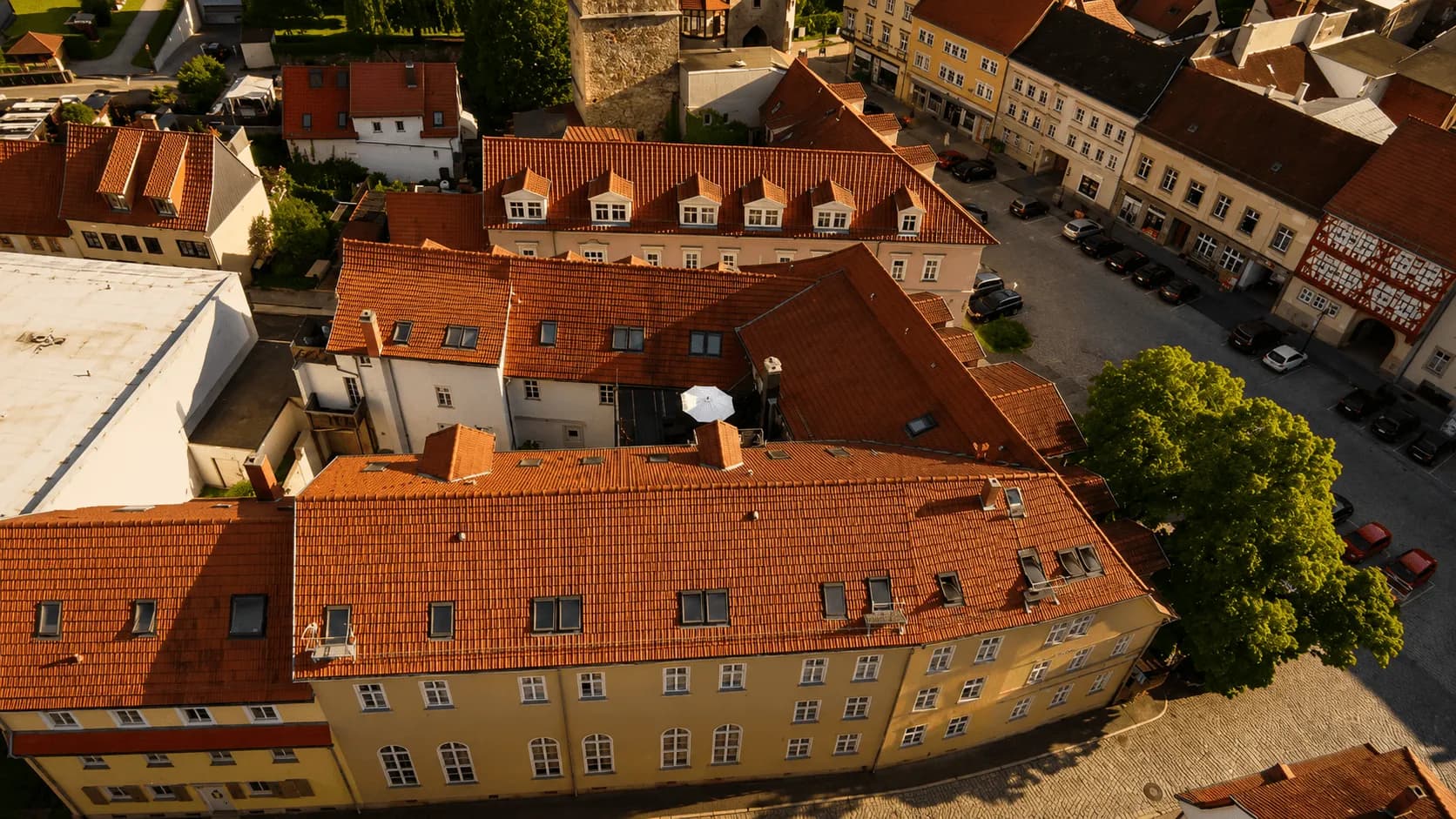 Draufsicht/Luftaufnahme des mehrflügeligen Gebäudeensembles im Abendlicht mit Ziegeldächern samt Dachgauben, befestigtem Innenhof mit Außenterrasse und Sonnenschirmen sowie Pkw-Stellplätzen.