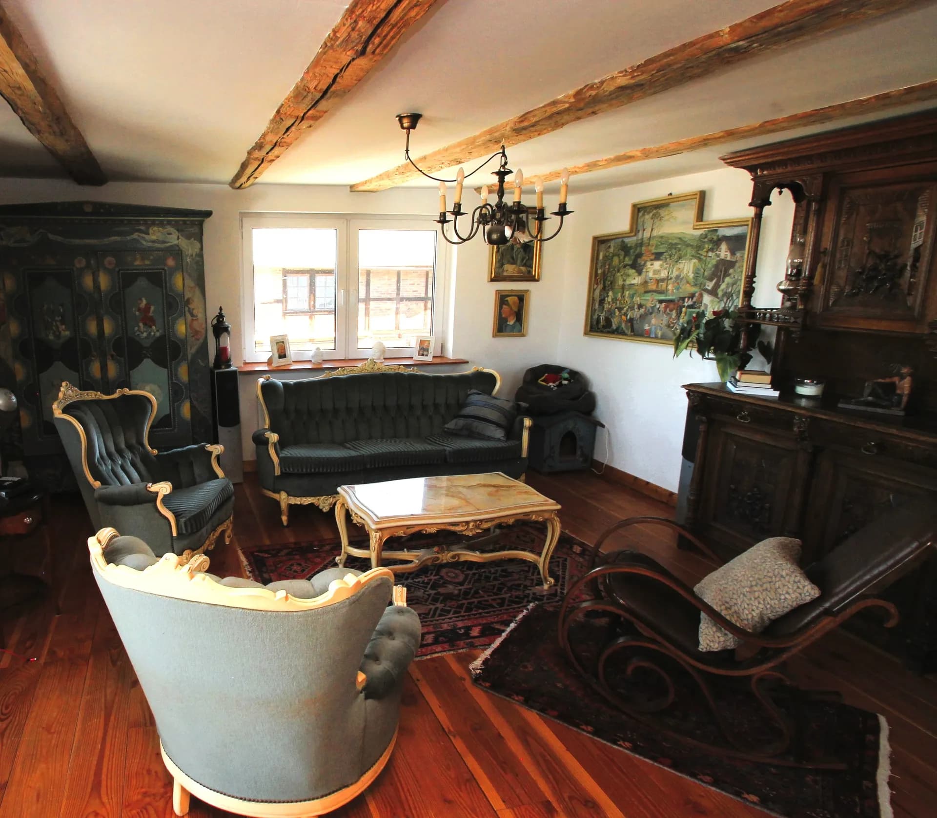 Gemütliches Wohnzimmer im klassischen Landhausstil mit sichtbaren Holzbalkendecken, antiken Polstermöbeln, Couchtisch, Schaukelstuhl und hochwertigem Holzfußboden.