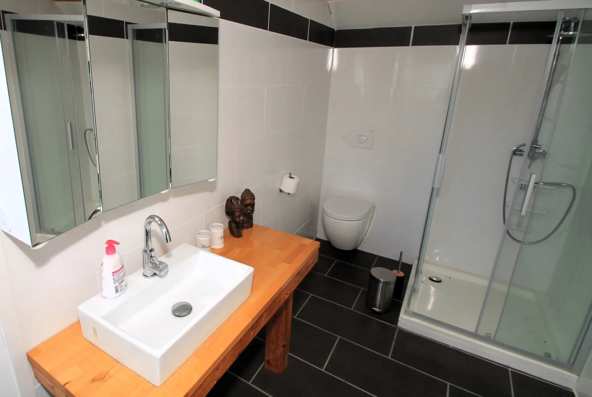 Modernes Duschbad mit dunklen Bodenfliesen, Glasduschkabine, wandhängendem WC, dreitürigem Spiegelschrank sowie massivem Holzwaschtisch mit rechteckigem Aufsatzwaschbecken.