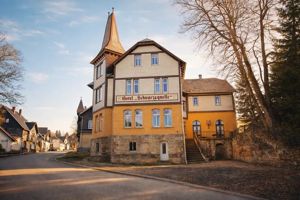 Repräsentative Straßenansicht des Hotels „Schwarzaquelle“ im warmen Sonnenlicht mit markantem Eckturmamt Spitzdach, Sandsteinsockel, Zierverschieferung und Außentreppe zur Terrasse.