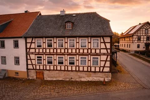 Atmosphärische Außenaufnahme des Fachwerkhauses im Abendlicht mit sichtbarem Holzfachwerk, Sandsteinsockel mit Kellerzugang und historischem Straßenpflaster.