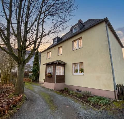 Perspektivische Außenansicht des Mehrfamilienhauses mit Zufahrt, holzverkleidetem Windfang, Dachgauben und natürlichem Baumbestand.