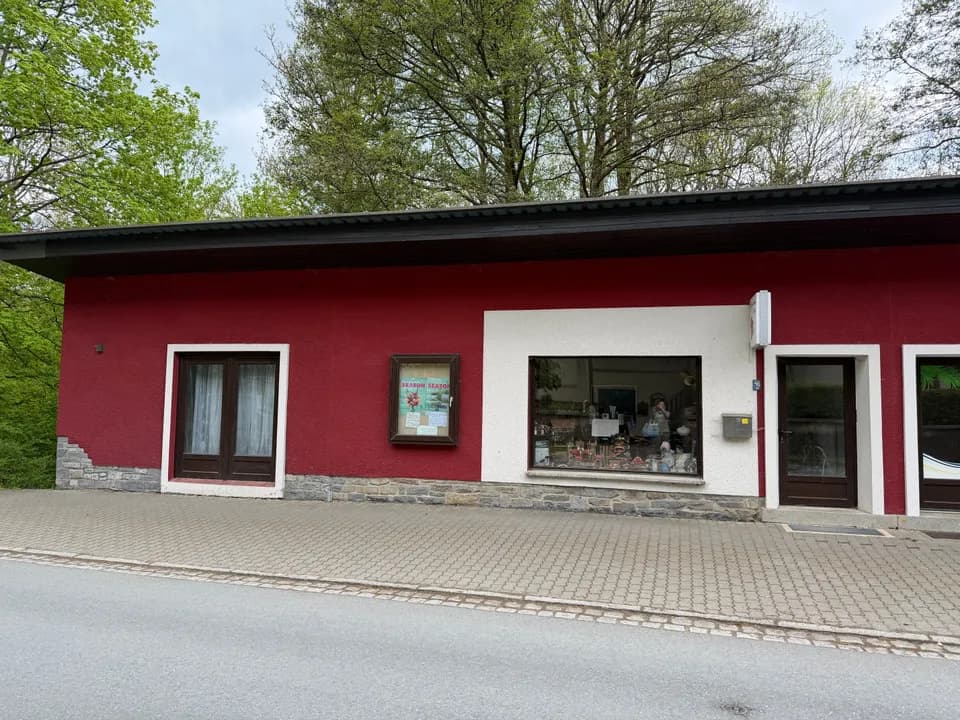 Außenansicht des freistehenden Gewerbe- oder Wohngebäudes in markantem Weinrot mit Natursteinsockel, Schaufensterfront und separatem Straßenzugang.