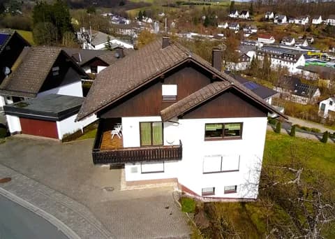 Schrägluftaufnahme des gepflegten Einfamilienhauses mit Giebel-Holzverschalung, großem Balkon, gepflastertem Hofraum, Garage und malerischem Ausblick über die Tallage.