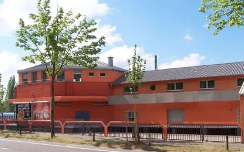 Straßenansicht der Gewerbe- und Gastronomieimmobilie in markantem Terrakotta-Orange mit polygonalem Kopfbau, Blechdach, Hallentrakt und eingefriedetem Vorplatz. Screenshot 2026-03-14 065023.jpg