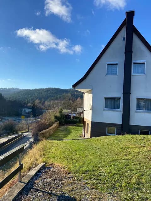 Seitenansicht des Wohnhauses mit ansteigendem Gartengrundstück, Außenkamin und idyllischem Fernblick auf Wald und Berge.