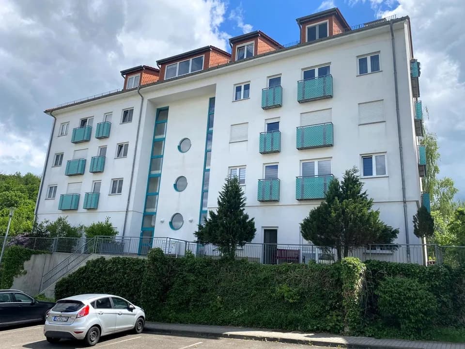 Außenansicht des modernen Mehrfamilienhauses mit türkisfarbenen französischen Balkonen, verglastem Treppenhausturm und Pkw-Stellplätzen.