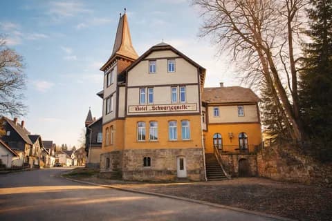 Repräsentative Straßenansicht des Hotels „Schwarzaquelle“ im warmen Sonnenlicht mit markantem Eckturmamt Spitzdach, Sandsteinsockel, Zierverschieferung und Außentreppe zur Terrasse.